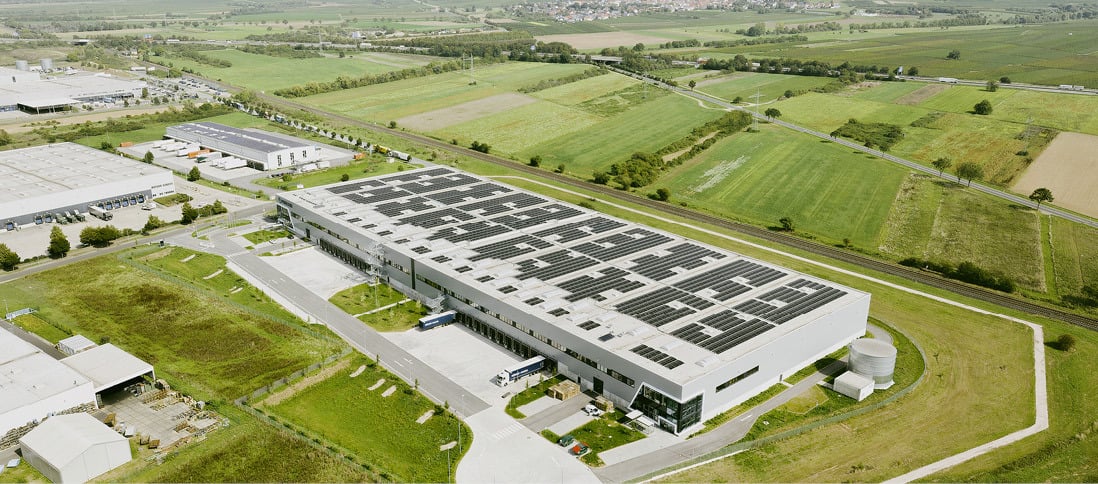 A large logistics facility with solar panels on the roof, surrounded by green fields.